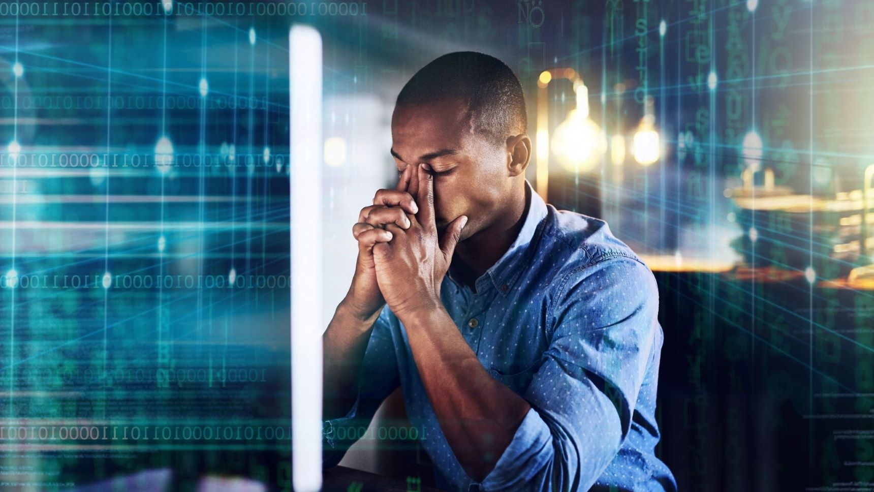 Stressed engineer pinches the bridge of his nose while sitting at a desk. computer code can be seen super imposed around the edges of the frame.